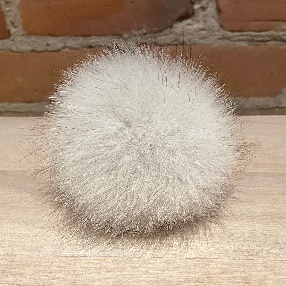 A fluffy pom-pom ball in a light cream or off-white fox fur, resting on a light wood surface with a brick wall background.