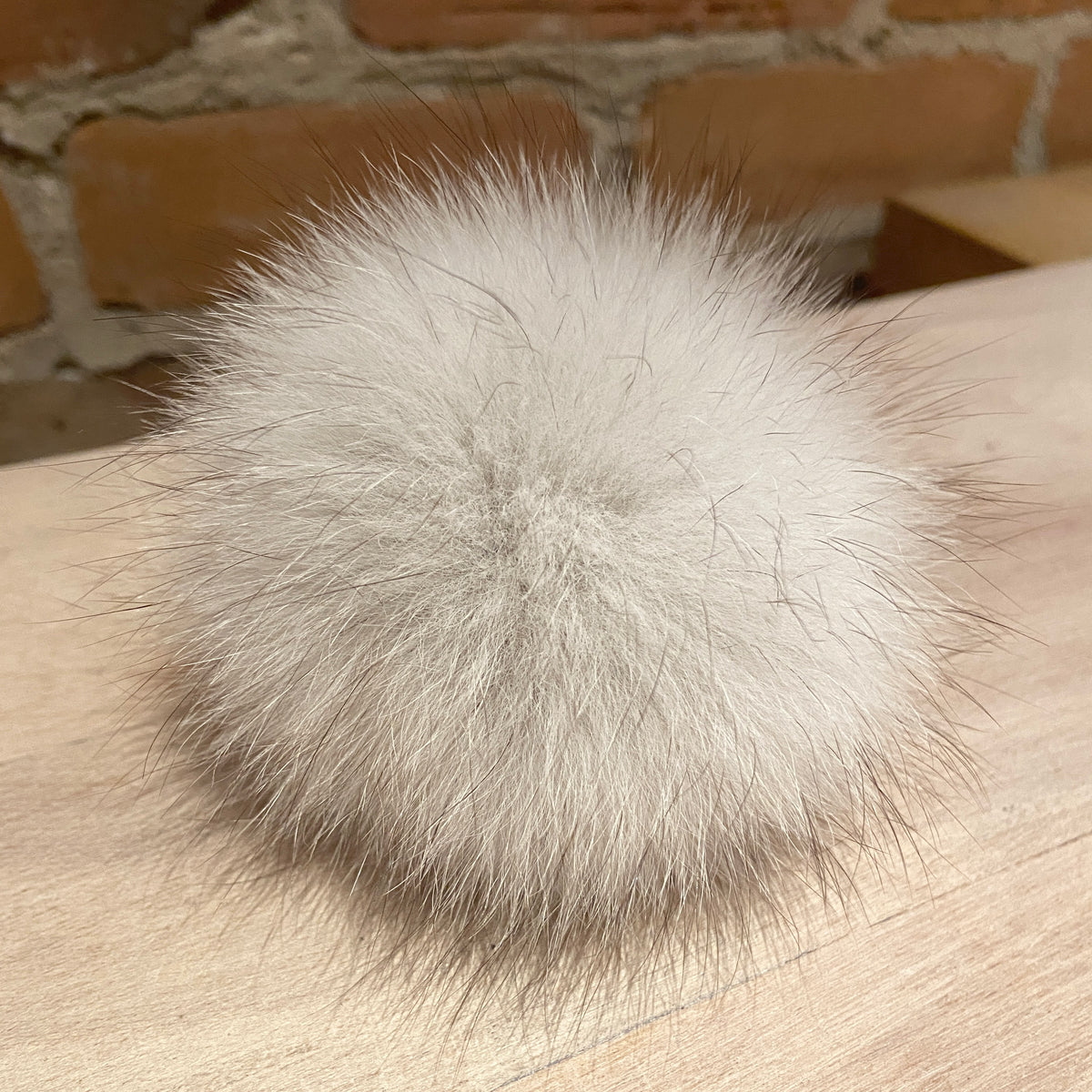 A fluffy pom-pom ball made of light gray and off-white fur, resting on a light wood surface with a brick wall in the background.