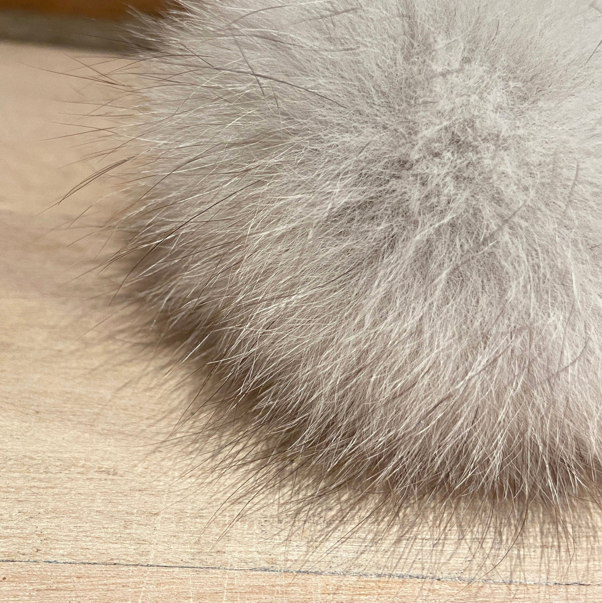 A close-up of a fluffy fox fur pom-pom in a light gray color resting on a light wood surface.