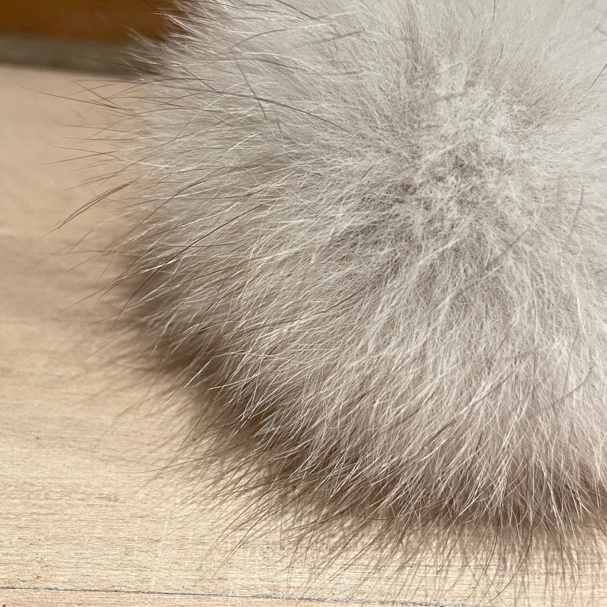 A close-up of a fluffy fox fur pom-pom in a light gray color resting on a light wood surface.