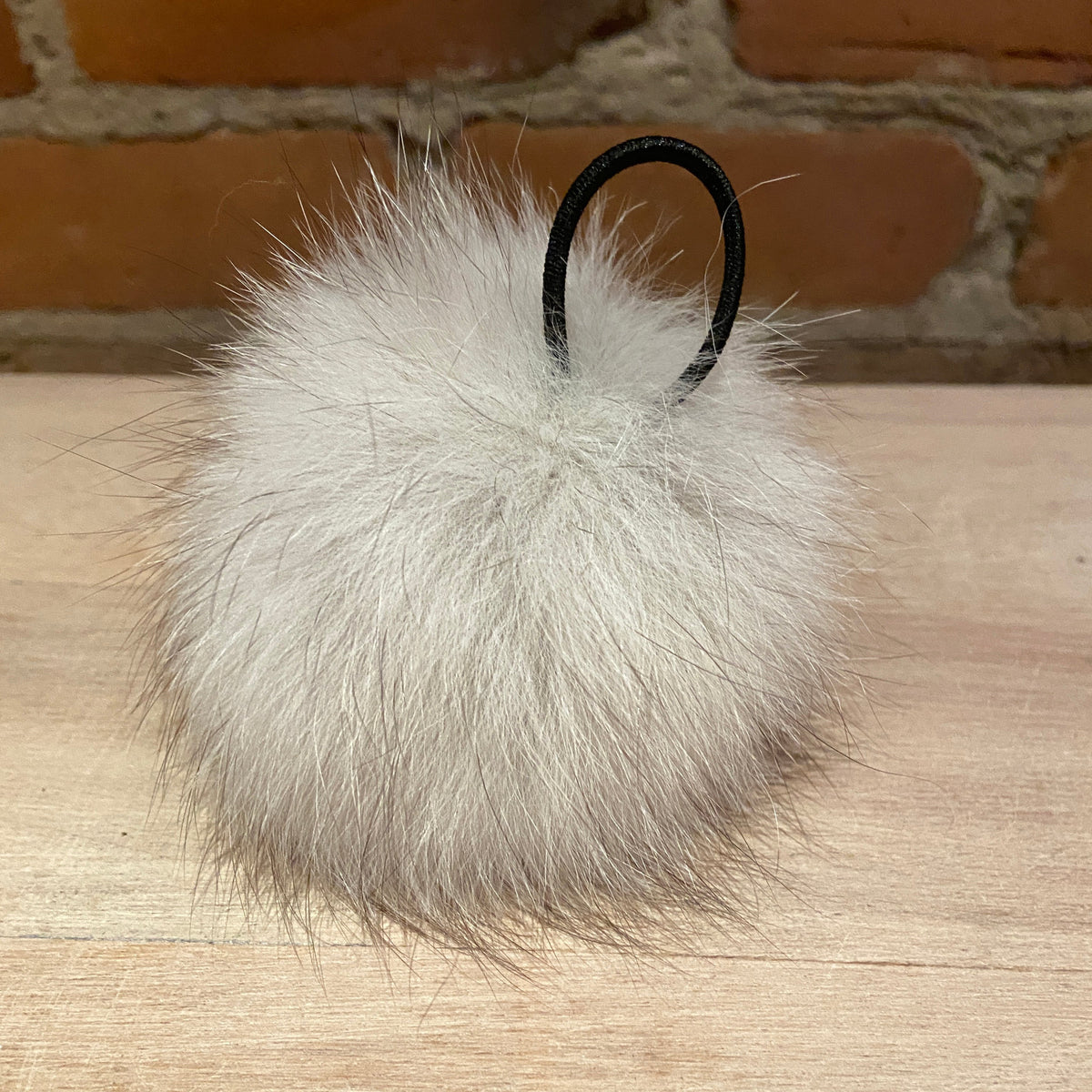 A fluffy white fox-fur pom-pom with a black elastic loop, resting on a light wood surface with a brick wall background.