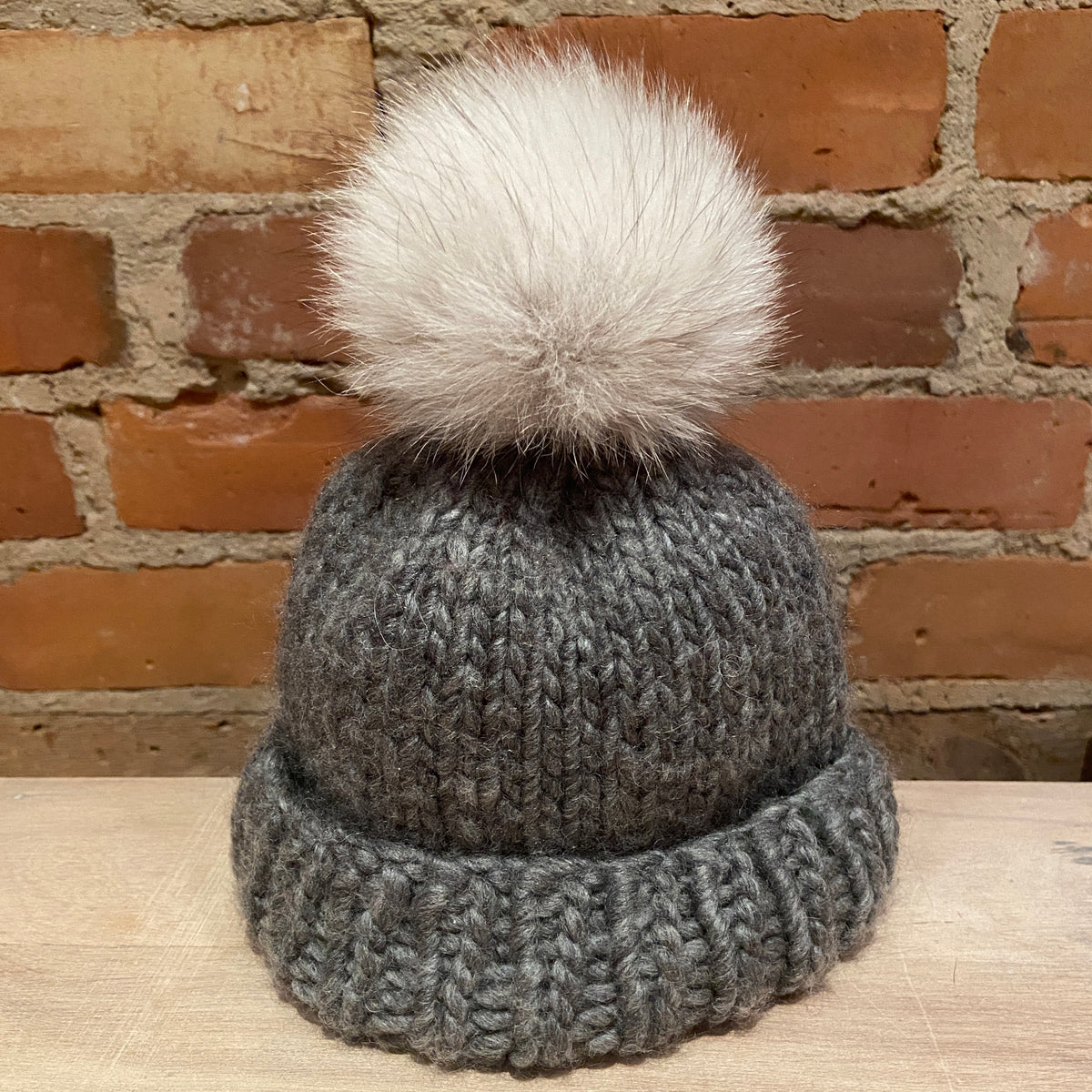 A small gray ribbed knit beanie with a small fluffy cream fox-fur pom-pom, displayed on a light wood surface against a rustic red-brown brick wall background.