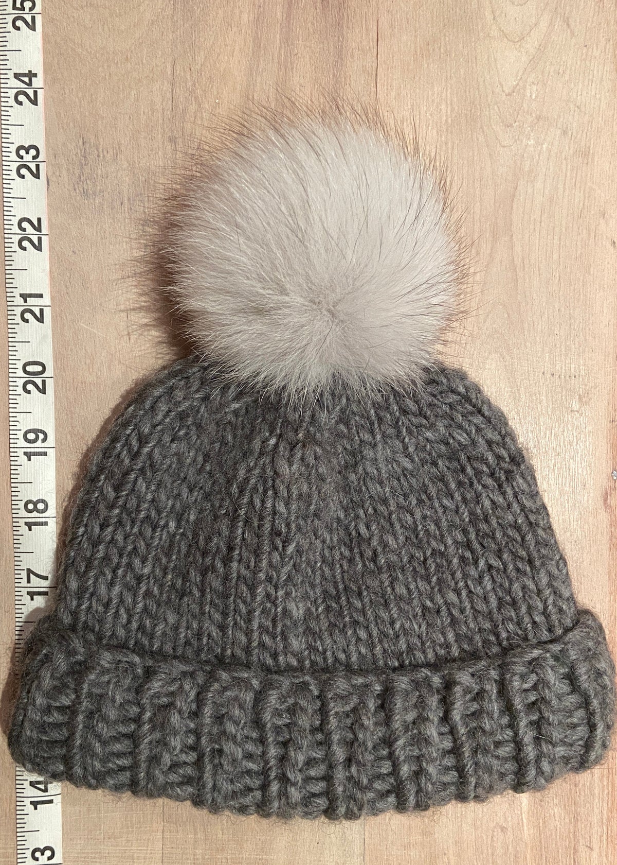 A small gray ribbed knit beanie with a small fluffy cream pom-pom, laid flat on a light wood surface next to a measuring tape ruler.