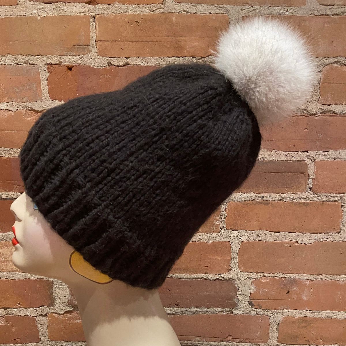 A black ribbed knit beanie hat with a fluffy white pom-pom, displayed on a mannequin head against a red brick wall background.
