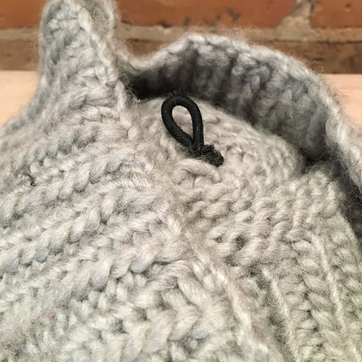 Close-up of a chunky knit light gray garment, showing the textured stitch pattern and a small black loop detail, with a brick wall in the background.