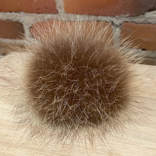 A brown raccoon fur pom-pom ball resting on a light wood surface with a brick wall in the background.