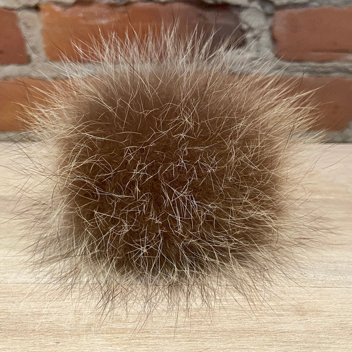 A round, fluffy brown raccoon fur ball with natural cream and gray tips, resting on a light wood surface with a brick wall background.