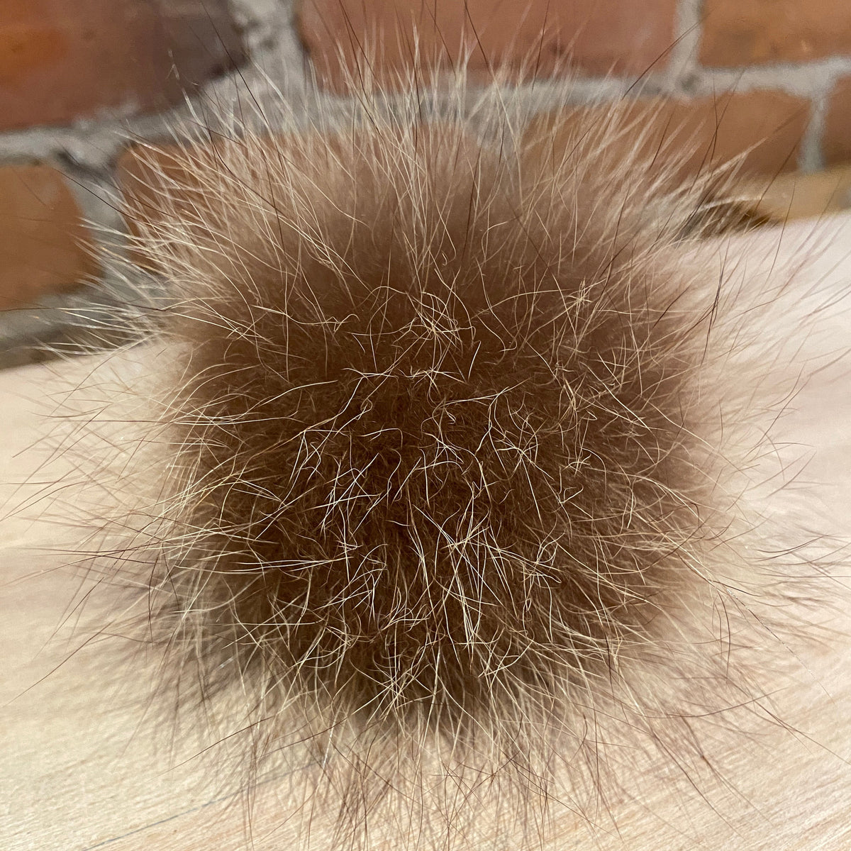A close-up of a fluffy brown raccoon fur pom-pom resting on a light wood surface with a blurred brick wall background.