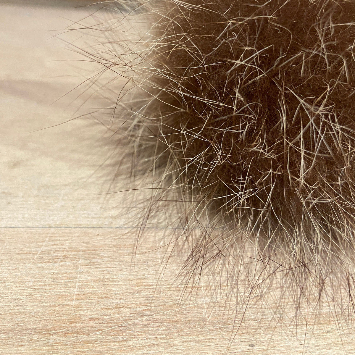 A close-up of a fluffy brown raccoon fur pom-pom resting on a light wood surface with a blurred brick wall in the background.