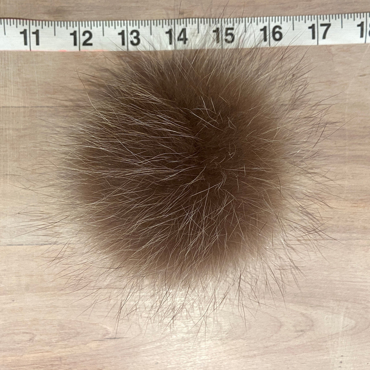 A round brown raccoon fur pom-pom with natural white tips, shown on a light wood surface with a measuring tape ruler at the top for scale.
