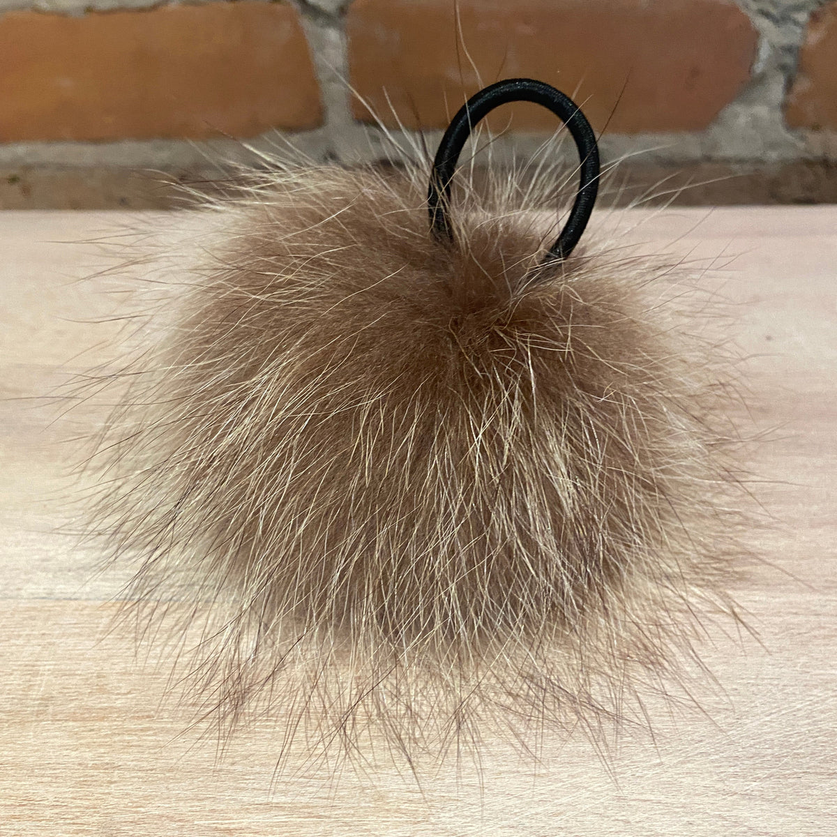 A fluffy raccoon pom pom in a light brown and beige tone, with a loop attachment, resting on a light wood surface with a brick wall in the background.