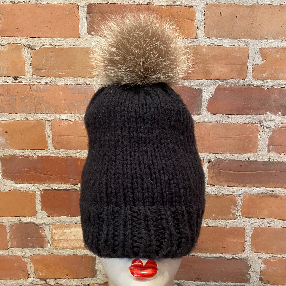 A black ribbed knit beanie with a raccoon fur pom-pom, displayed on a mannequin head with red lips in front of a brick wall background.