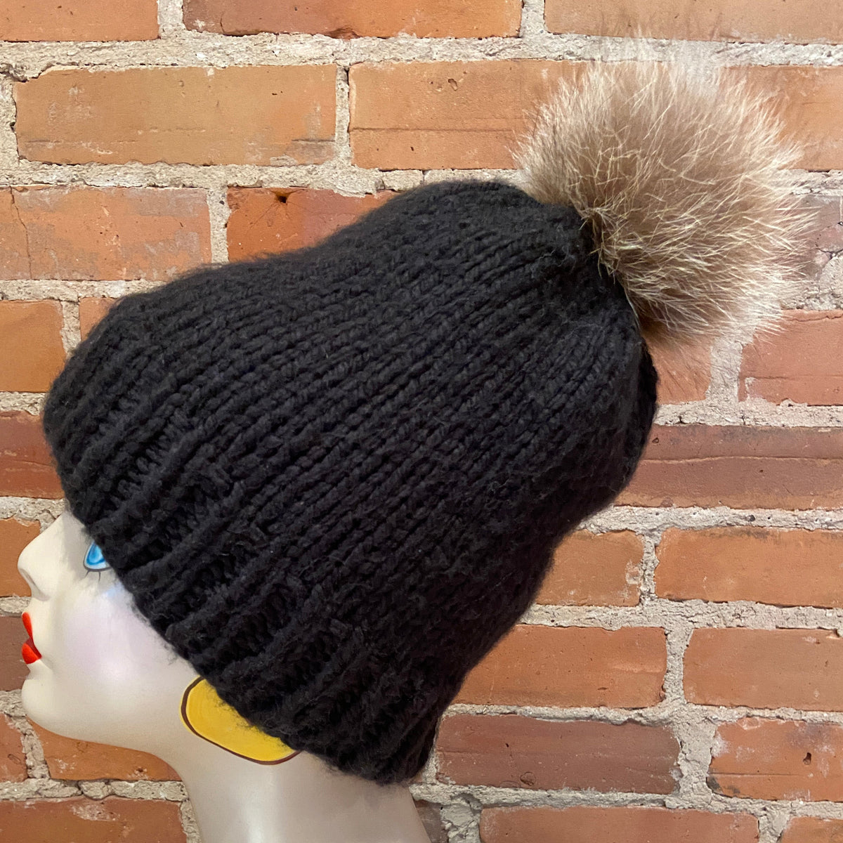 A black ribbed knit beanie with a fluffy brown and beige raccoon fur pom-pom, displayed on a mannequin head against a red brick wall background.