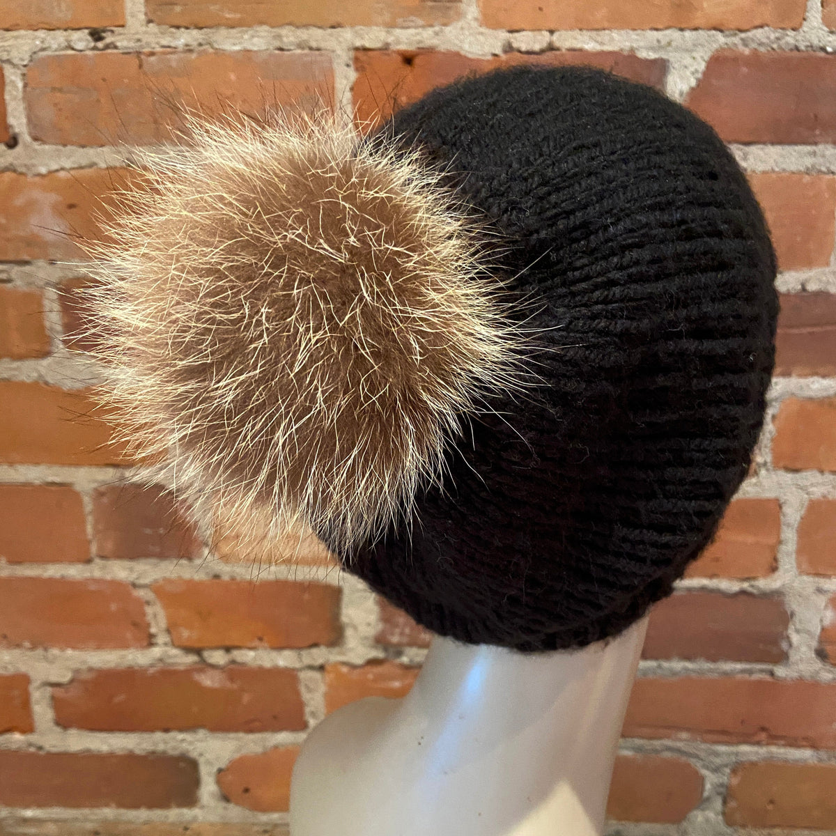 A black ribbed knit beanie hat with a fluffy tan raccoon-fur pom-pom, displayed on a white mannequin head against a red brick wall background.