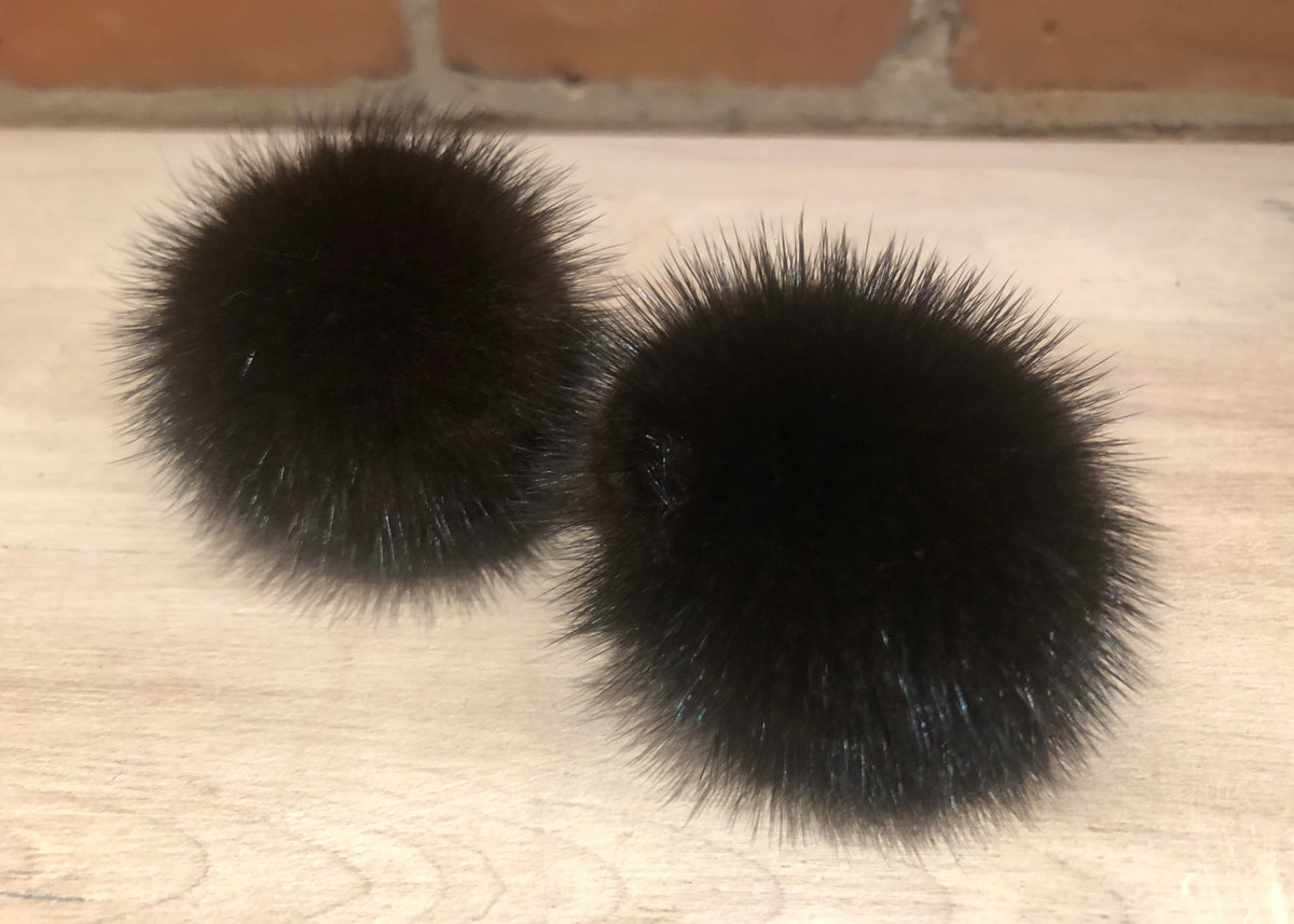 Two round black mink fur pom-poms resting on a light wood surface with a brick wall in the background.