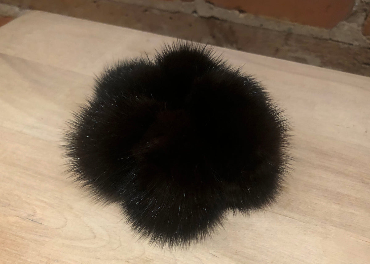 A black fluffy mink fur pom-pom accessory resting on a light-colored wooden surface with a brick wall in the background.