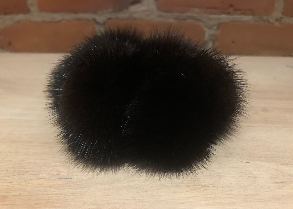 A black mink fur pom-pom accessory resting on a light wood surface with a brick wall background.