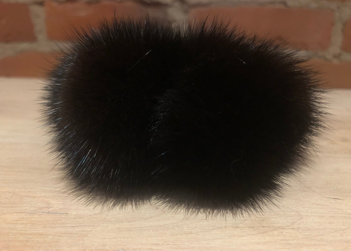 A pair of black mink fur pom-pom scrunchie resting on a light wood surface with a brick wall background.