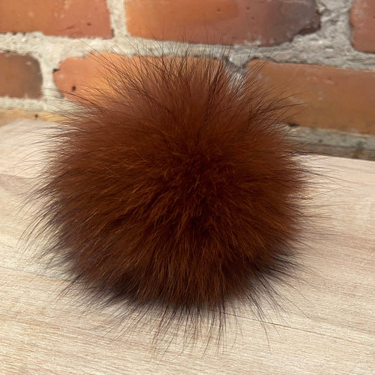 A fluffy, spherical burgundy fur pom-pom resting on a light-colored wooden surface with a brick wall background.