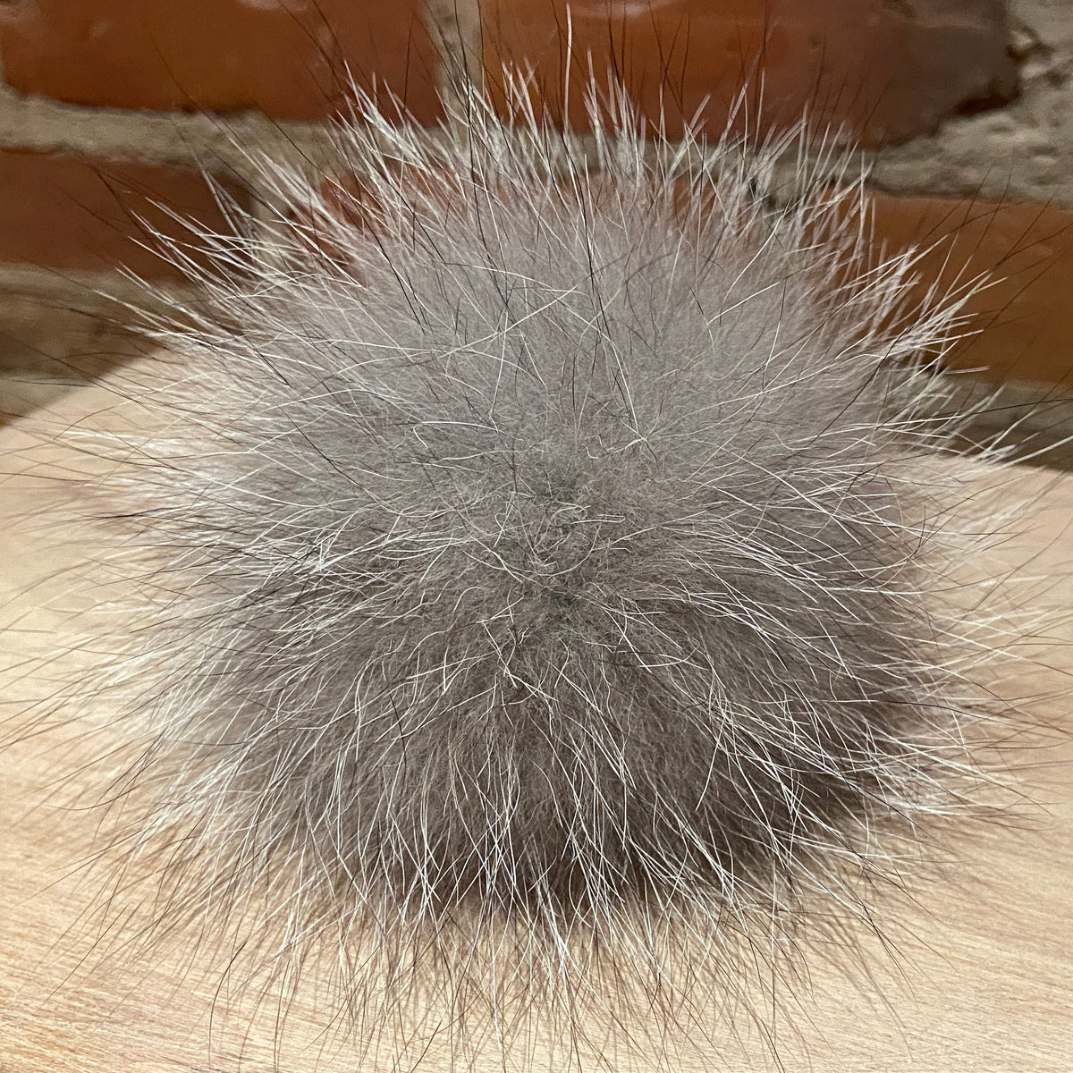 A fluffy gray fox fur pom-pom ball resting on a light wood surface with a brick wall background.
