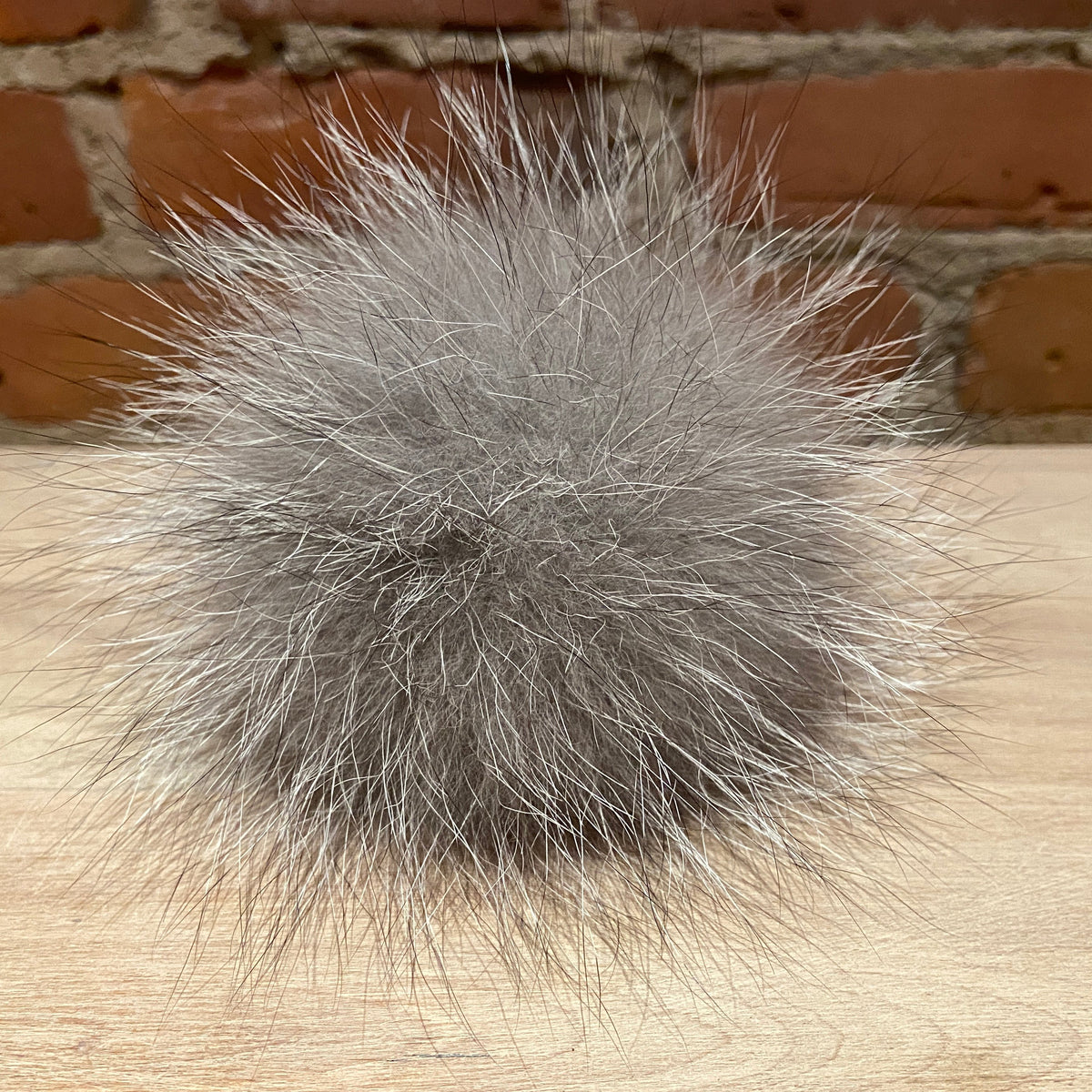 A fluffy gray fox  fur pom-pom ball resting on a light wood surface with a brick wall background.