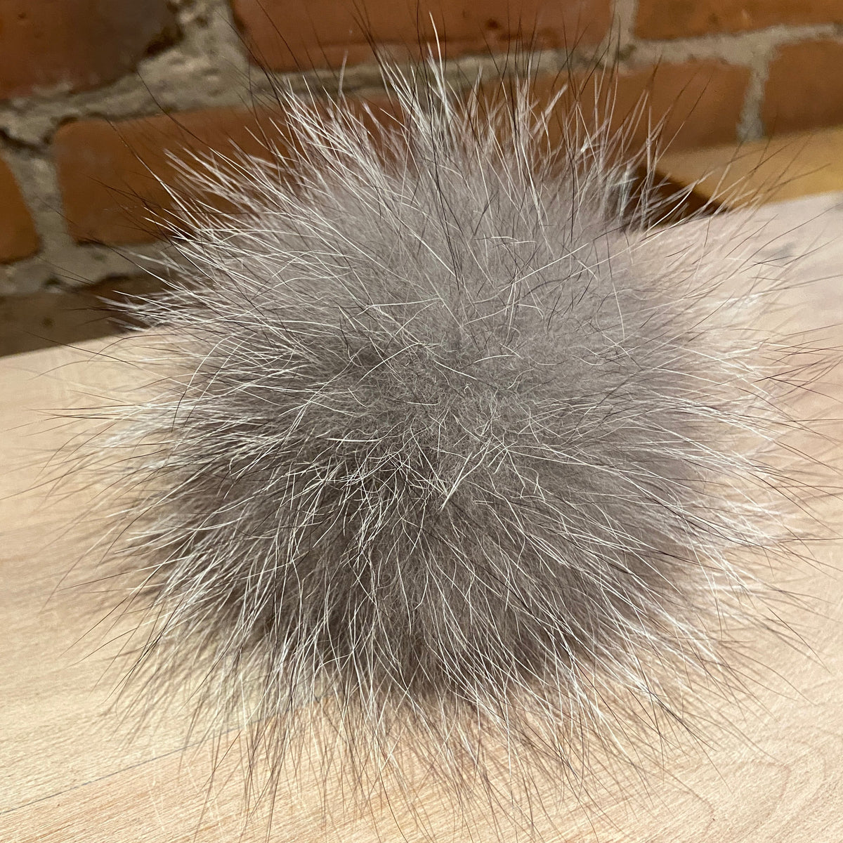 A fluffy gray fox-fur pom-pom accessory resting on a light wood surface with a brick wall in the background.
