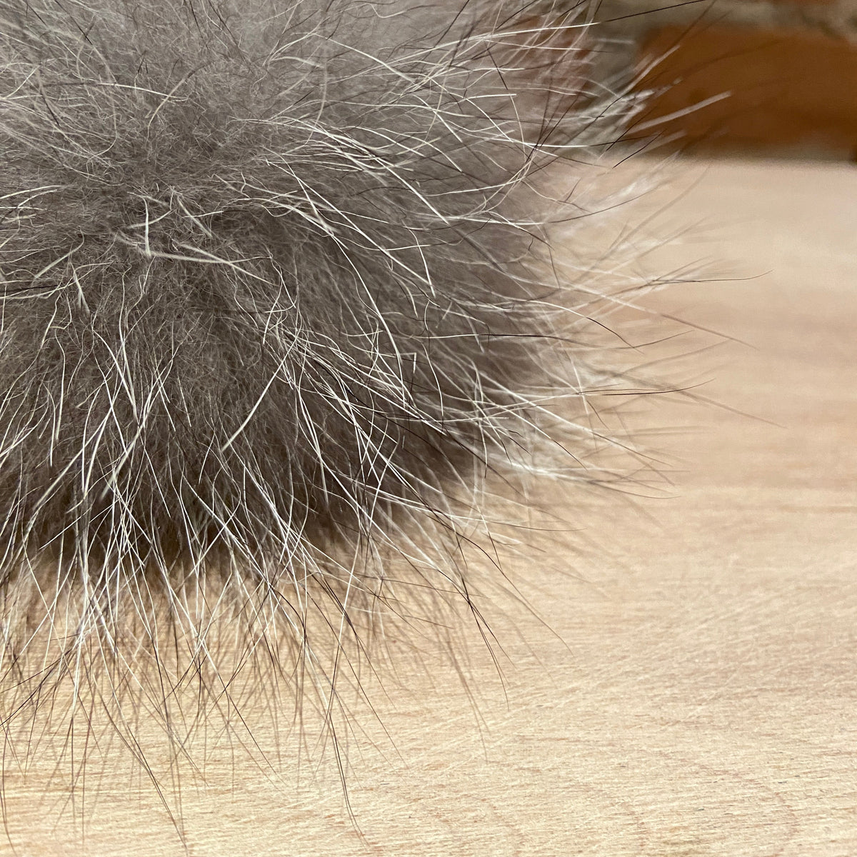 A close-up of a fluffy gray fox fur pom-pom with silver-gray tips, resting on a light wood surface with a blurred brick wall background.