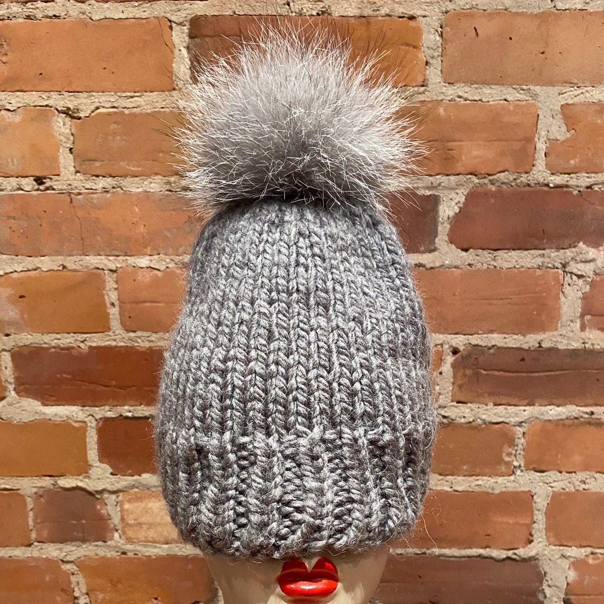 A gray chunky knit beanie with a fluffy gray fox fur pom-pom, displayed on a mannequin head with red lips in front of a red brick wall.