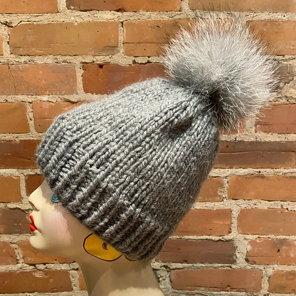 A gray ribbed knit beanie with a fluffy gray pom-pom, displayed on a mannequin head against a red-brown brick wall background.