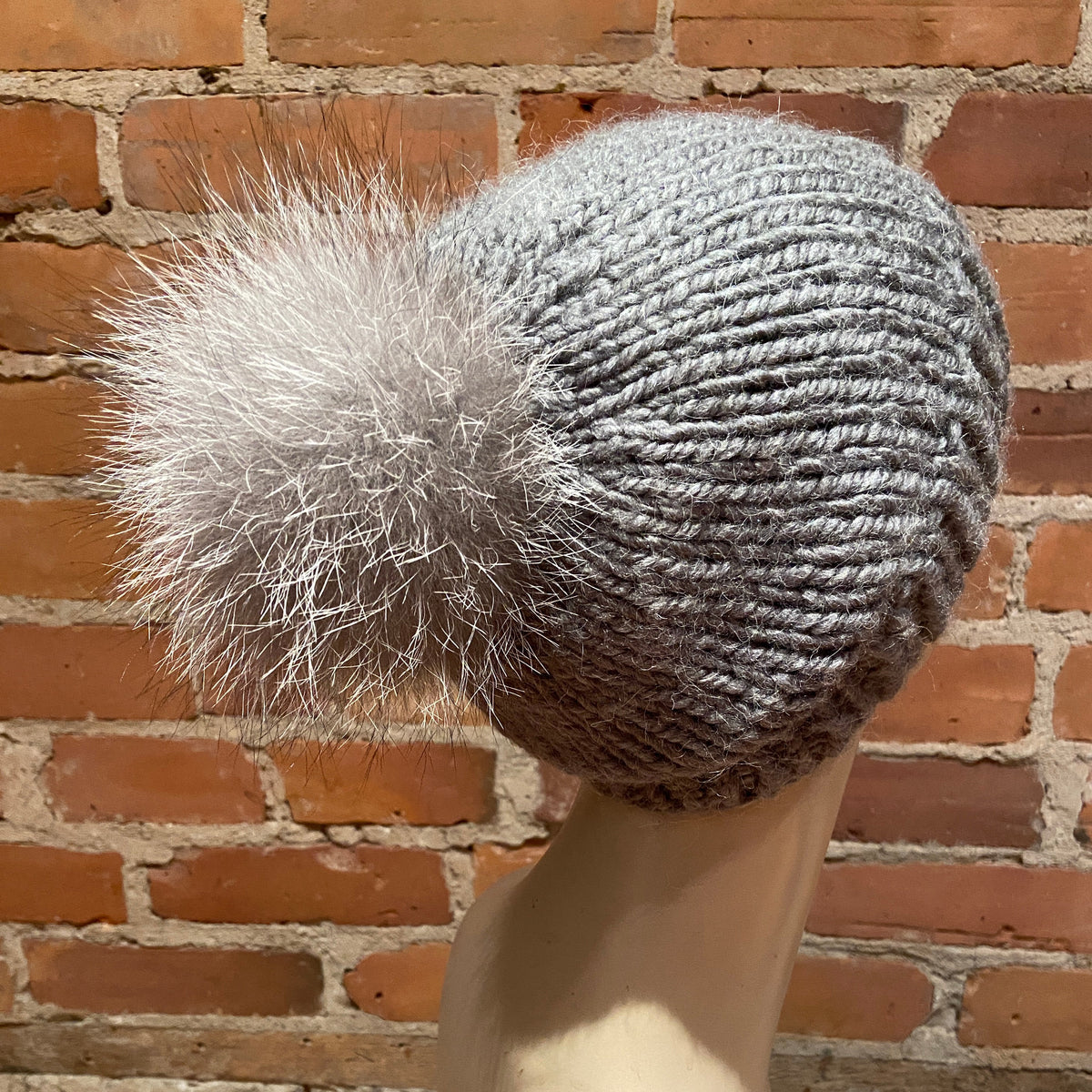 A gray ribbed knit beanie with a fluffy fox-fur pom-pom, displayed on a mannequin head against a red brick wall background.