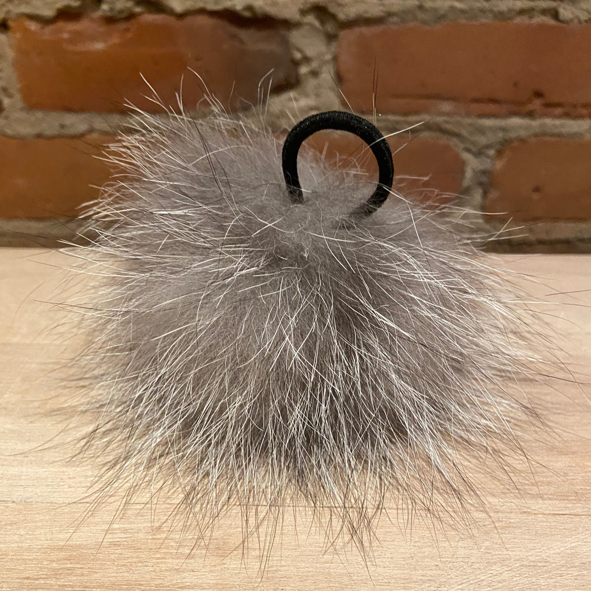A fluffy gray fox-fur pom-pom with a black elastic loop, resting on a light wood surface with a brick wall background.