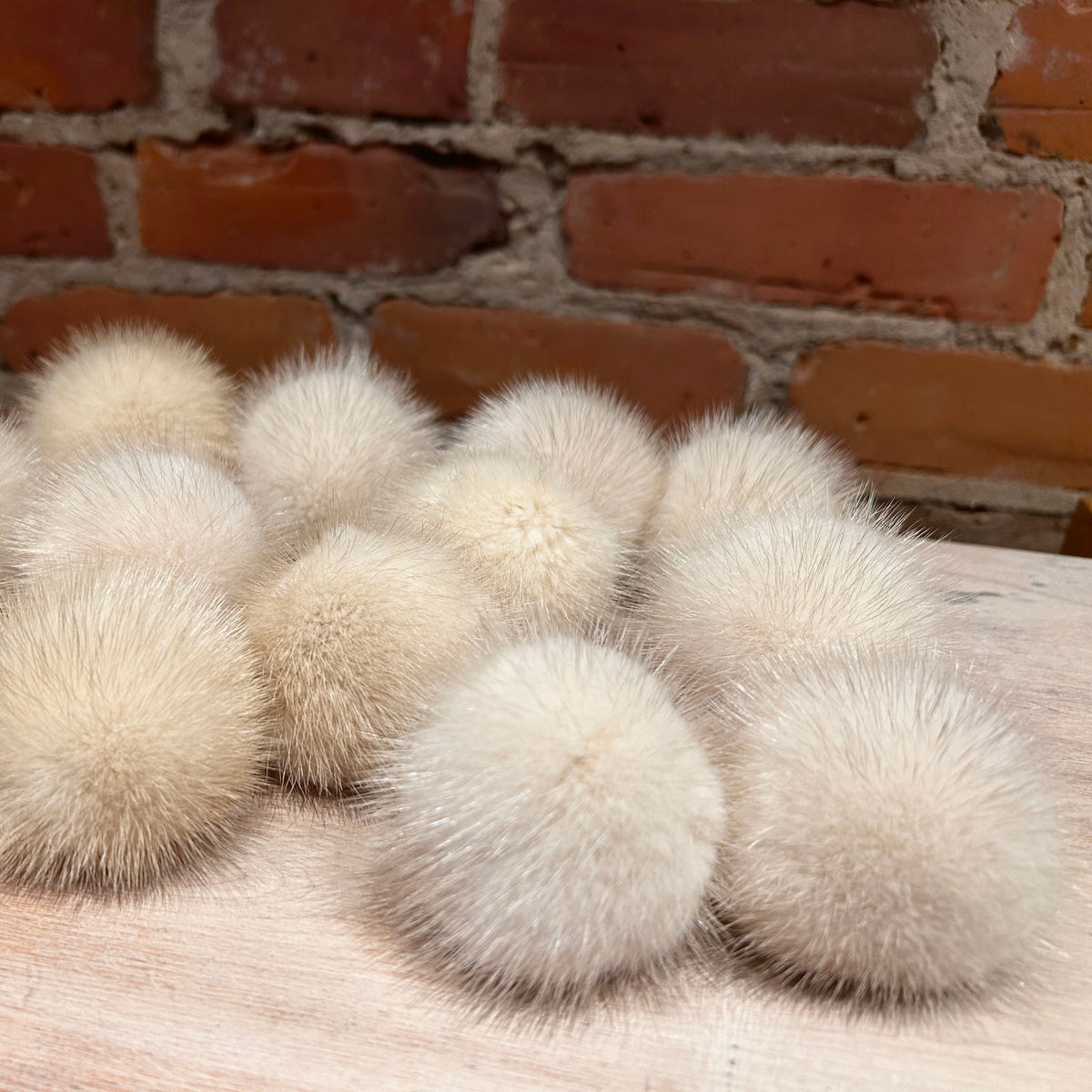 A group of cream-colored Mink Mini fur pom poms arranged on a light wooden surface in front of a red brick wall background.
