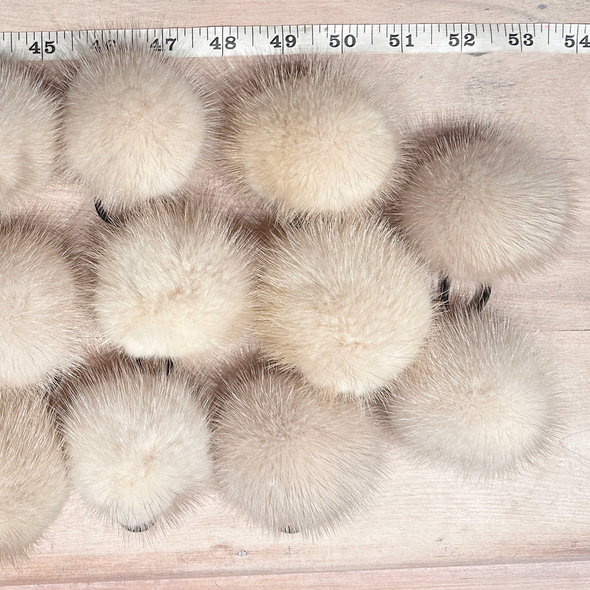 A set of fluffy mink mini -fur pom-poms in light beige and cream shades, arranged in rows on a light wood surface with a measuring tape visible at the top for scale.