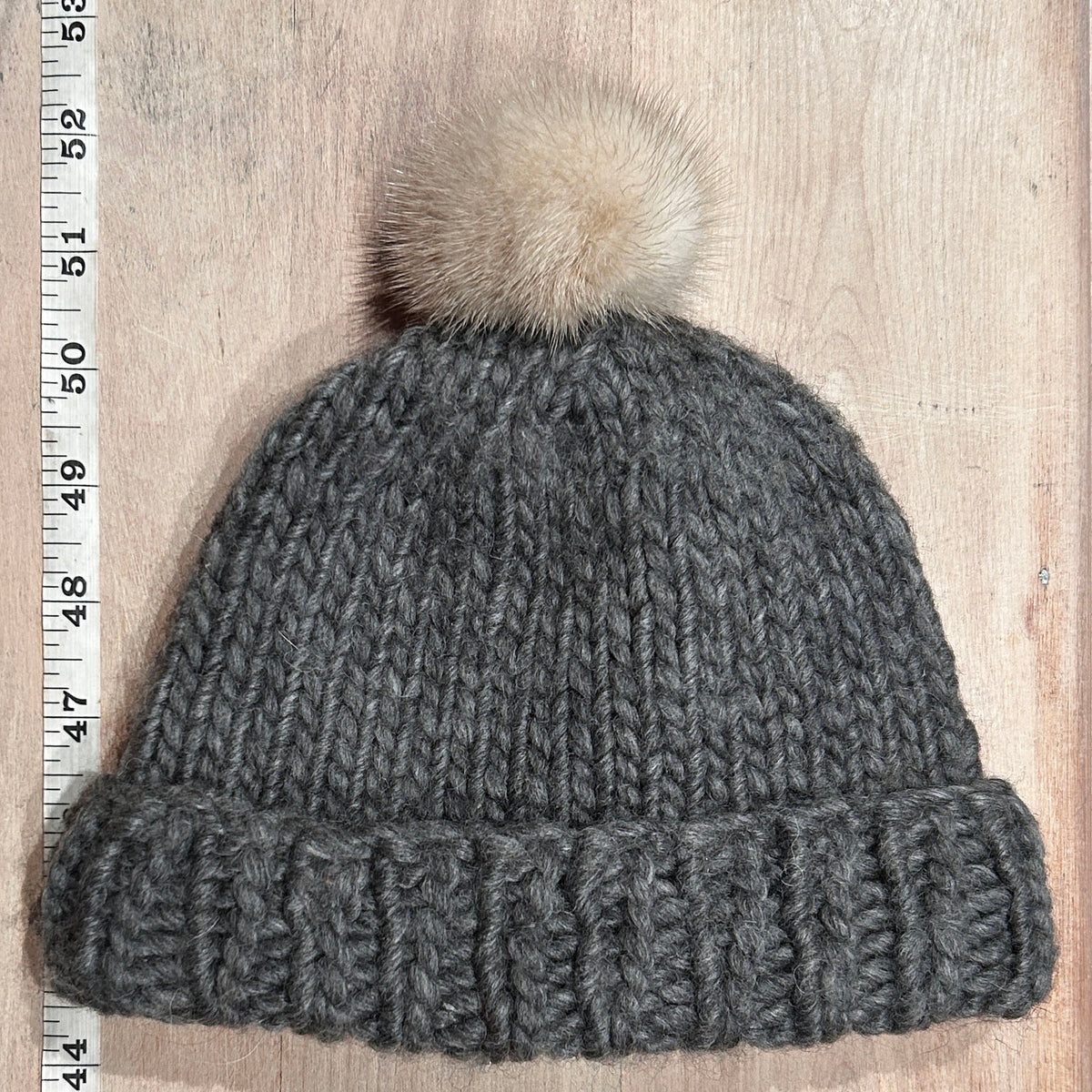 A dark gray chunky knit beanie with a fluffy cream mink mini pom-pom, laid flat on a light wood background next to a measuring ruler.