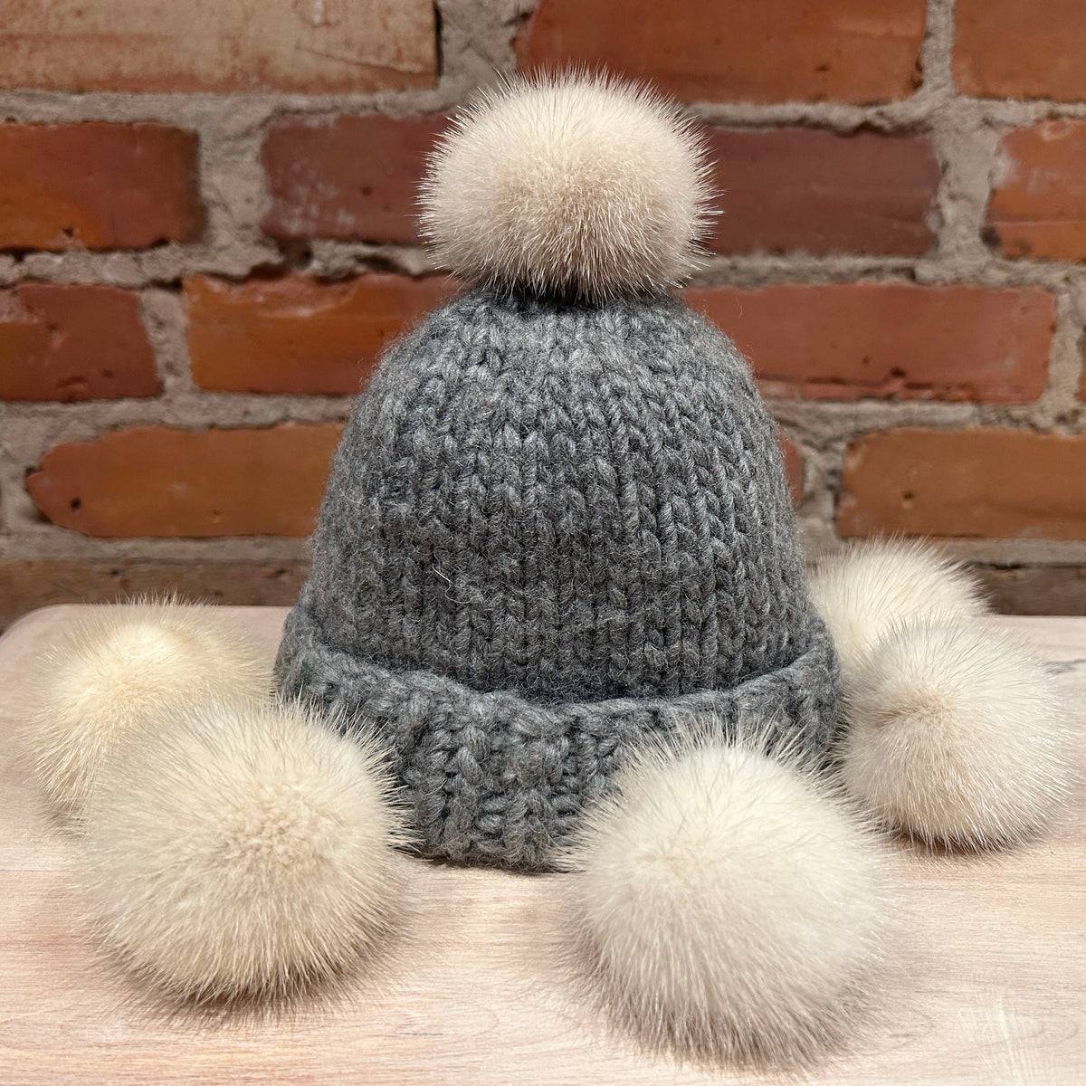 A gray knit pom-pom hat with four fluffy cream mink mini-fur pom-poms attached to the brim, displayed on a wooden surface in front of a brick wall.
