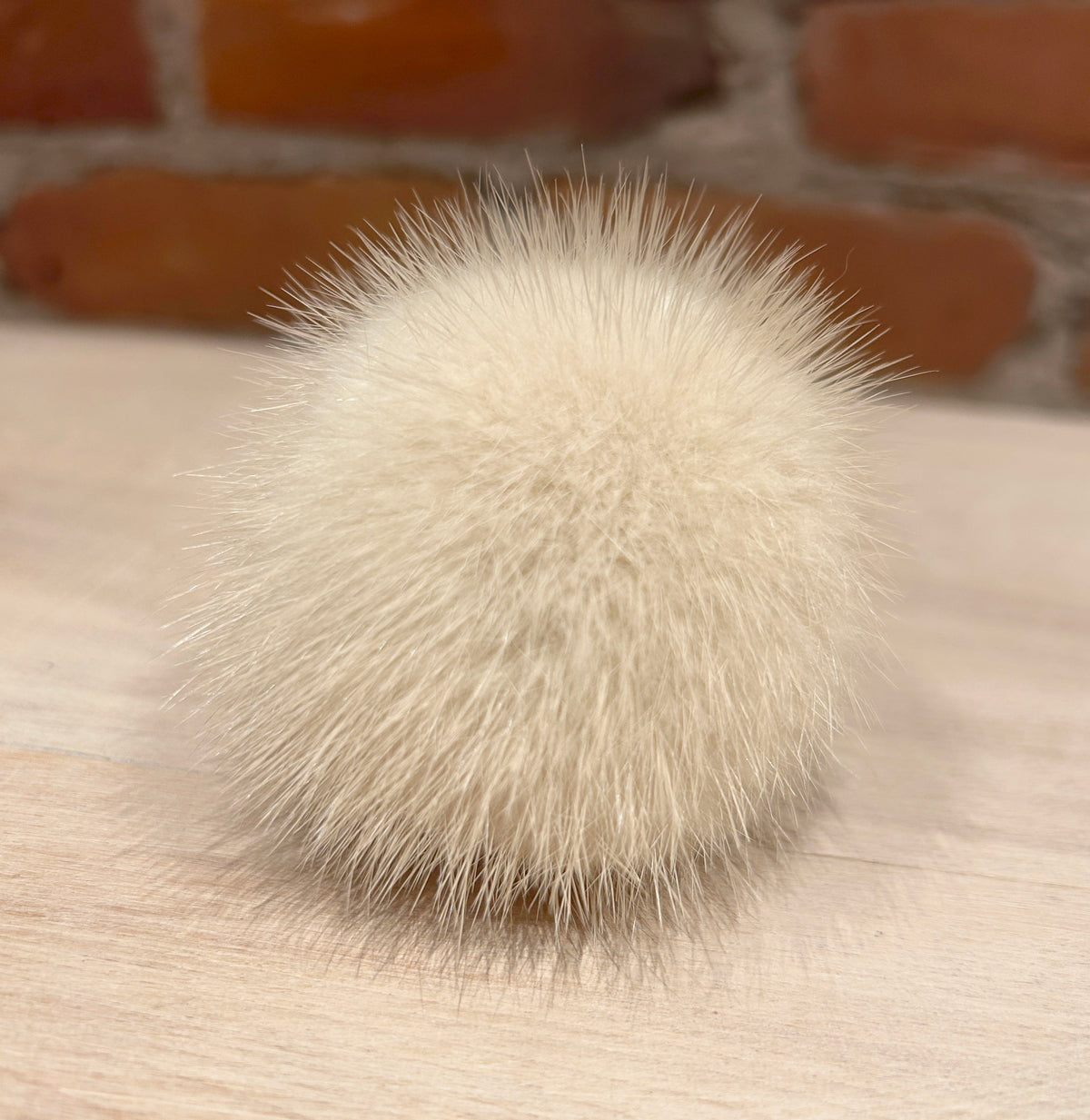 A fluffy cream-colored mink mini fur pom-pom accessory resting on a light wood surface with a blurred brick wall background.