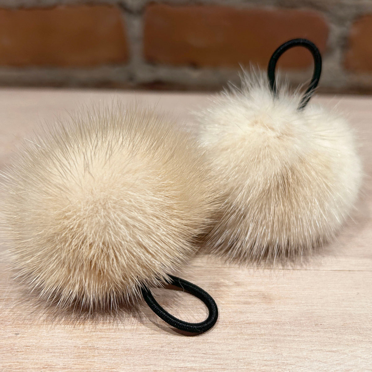 Two fluffy cream mink mini -fur pom-poms with black elastic loops, resting on a light wood surface with a brick wall background.