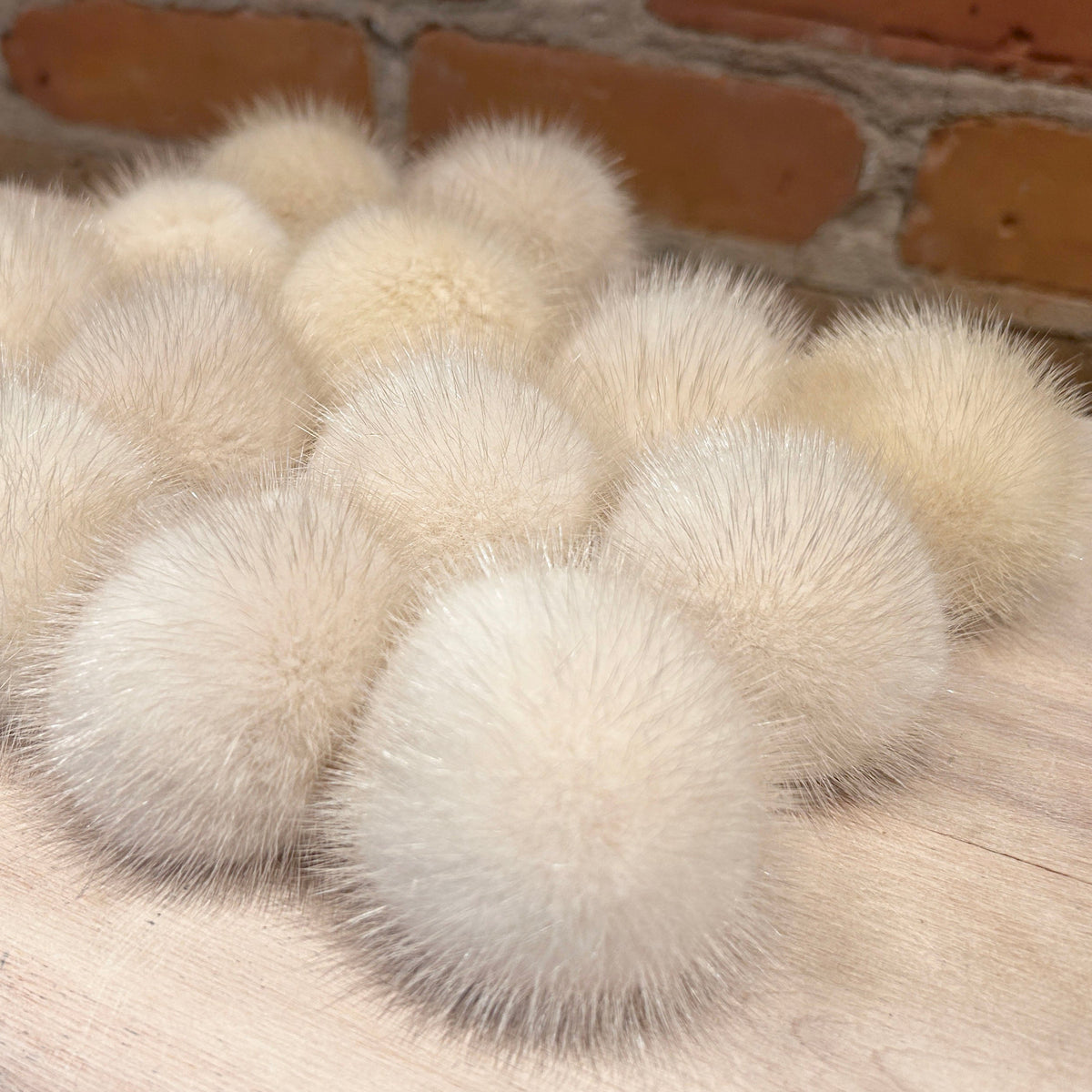A group of fluffy cream-colored pom-poms arranged on a light wood surface with a brick wall in the background.