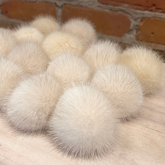 A group of fluffy cream-colored pom-poms arranged on a light wood surface with a brick wall in the background.