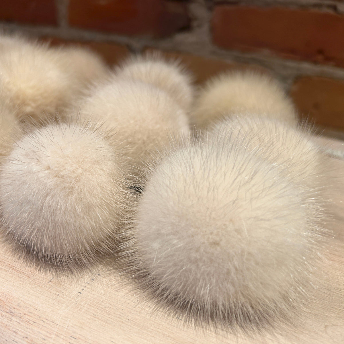 Several fluffy cream-colored mink mini fur pom poms arranged on a light wood surface with a blurred brick wall background.
