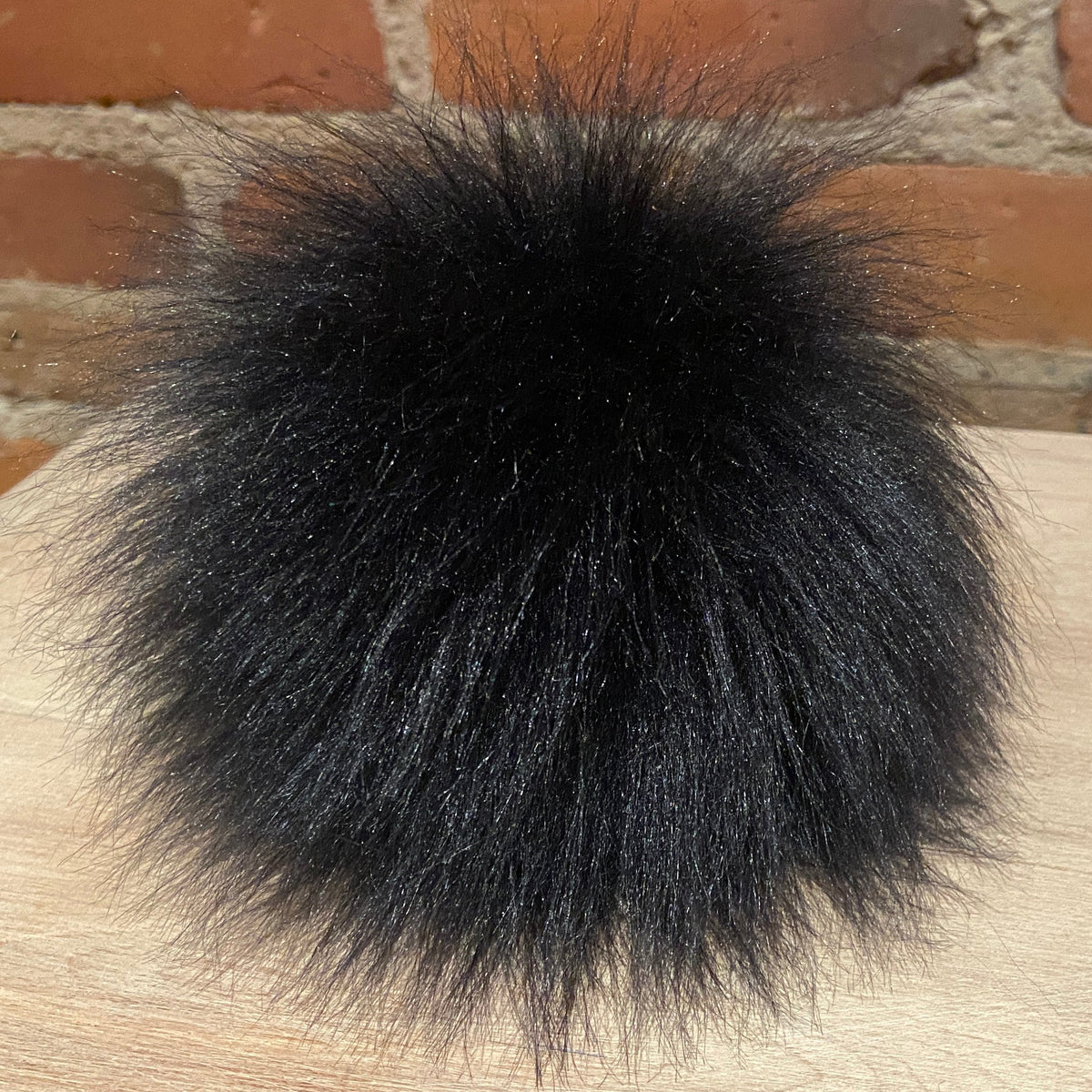 A fluffy black faux fur pom-pom resting on a light wood surface with a brick wall background.