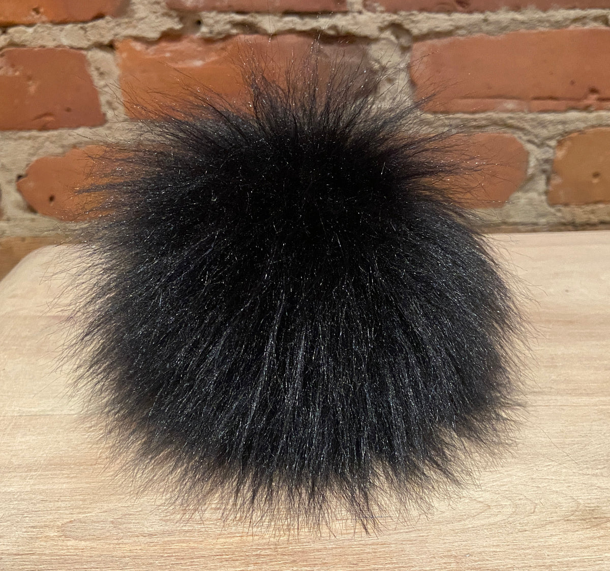 A fluffy black faux fur pom-pom accessory resting on a light wood surface with a brick wall background.