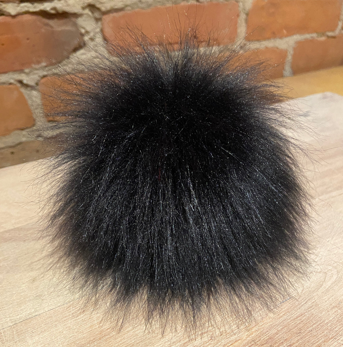 A fluffy black faux fur pom-pom accessory resting on a light wood surface with a brick wall background.