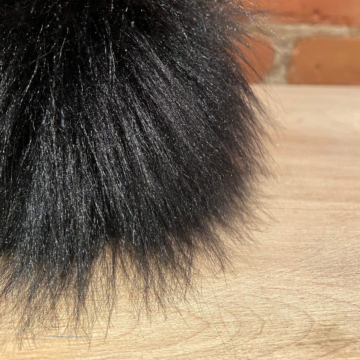 A close-up of a fluffy black faux fur pom-pom resting on a light wood surface with a blurred brick wall background.