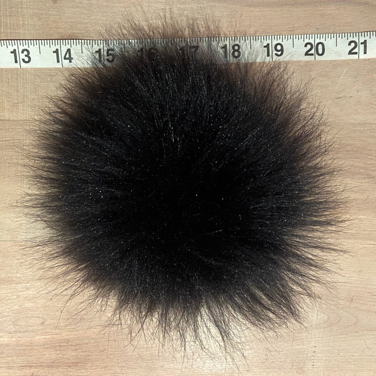 A round, fluffy black faux fur pom-pom shown on a light wood surface with a measuring tape ruler at the top for scale.