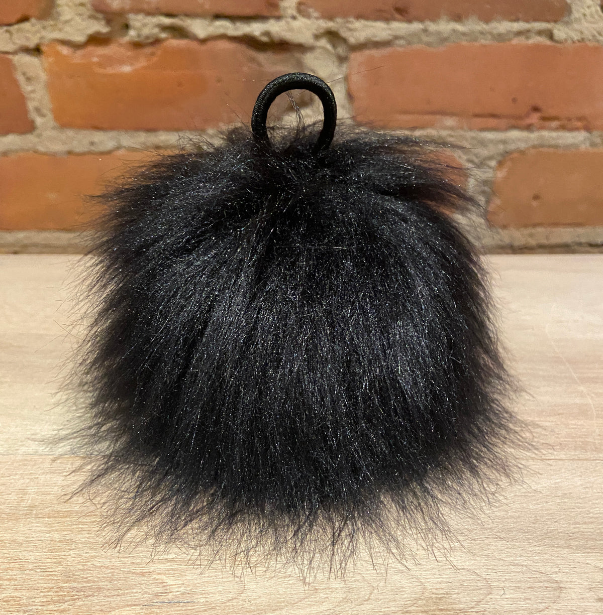 A fluffy black faux fur pom-pom bag charm with a black elastic loop, resting on a light wood surface in front of a brick wall background.