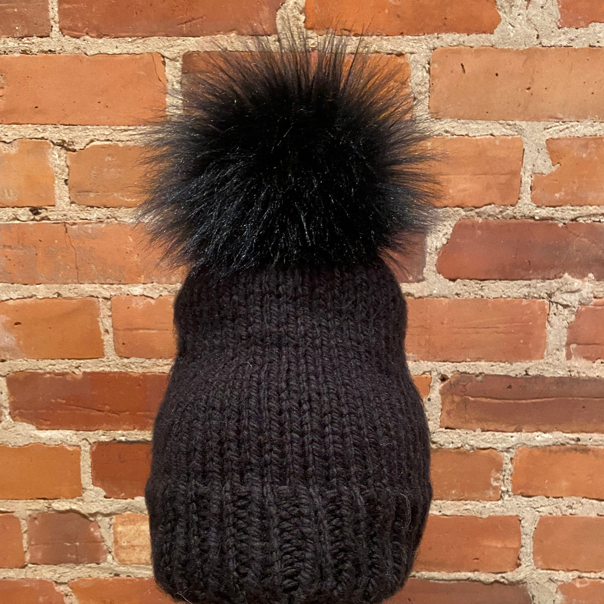 A black ribbed knit beanie with a large fluffy black pom-pom, displayed on a mannequin head with red lipstick in front of a red brick wall.