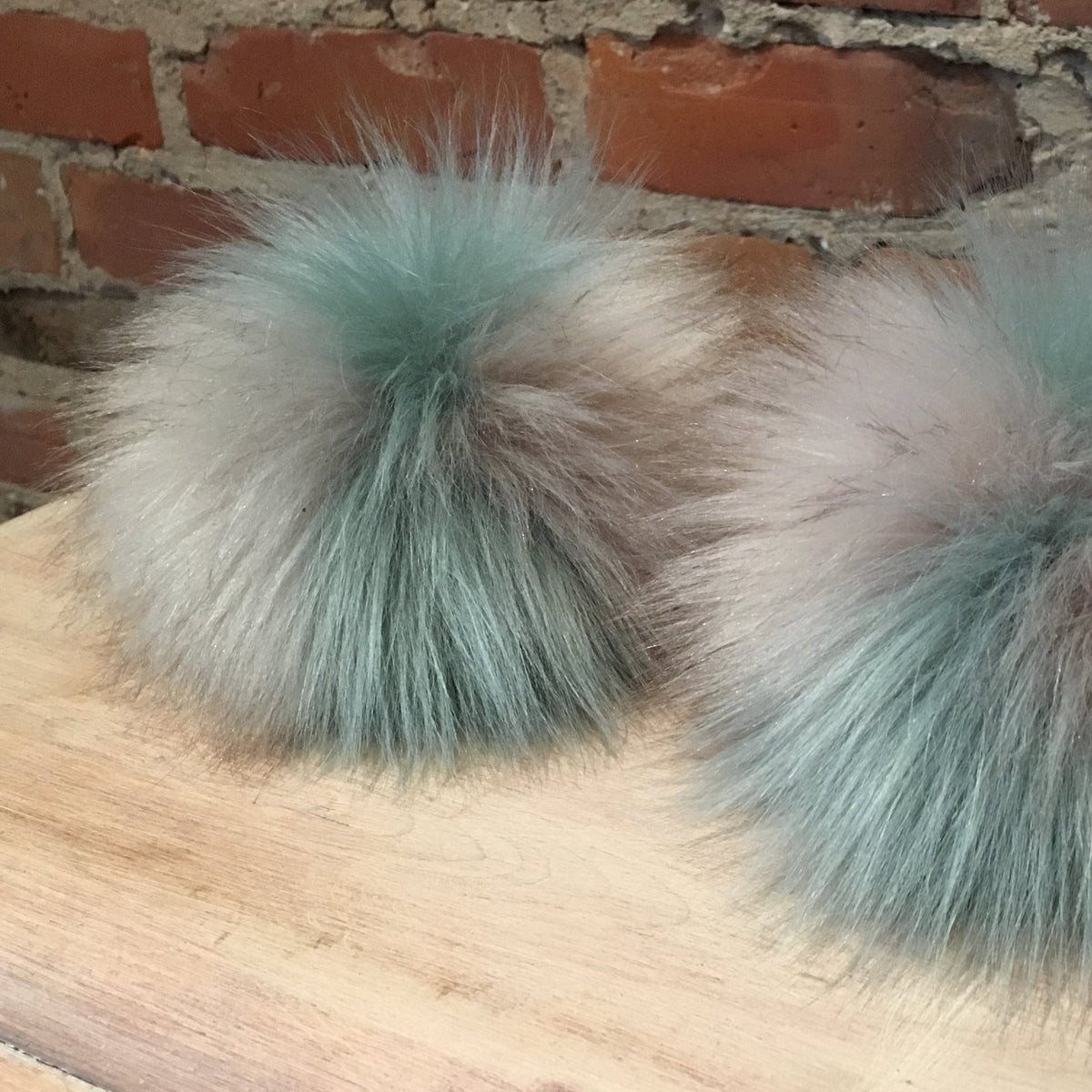 Two fluffy faux-fur pom-poms in a dusty sage green and taupe color, displayed on a wooden surface against a brick wall background.
