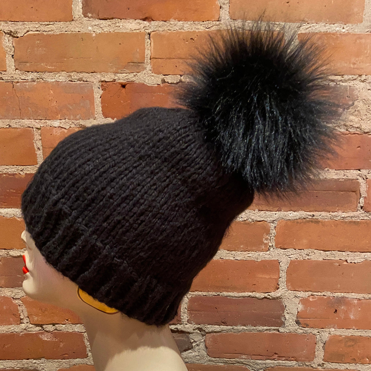 A black ribbed knit beanie with a large black faux fur pom-pom, displayed on a mannequin head against a red brick wall background.
