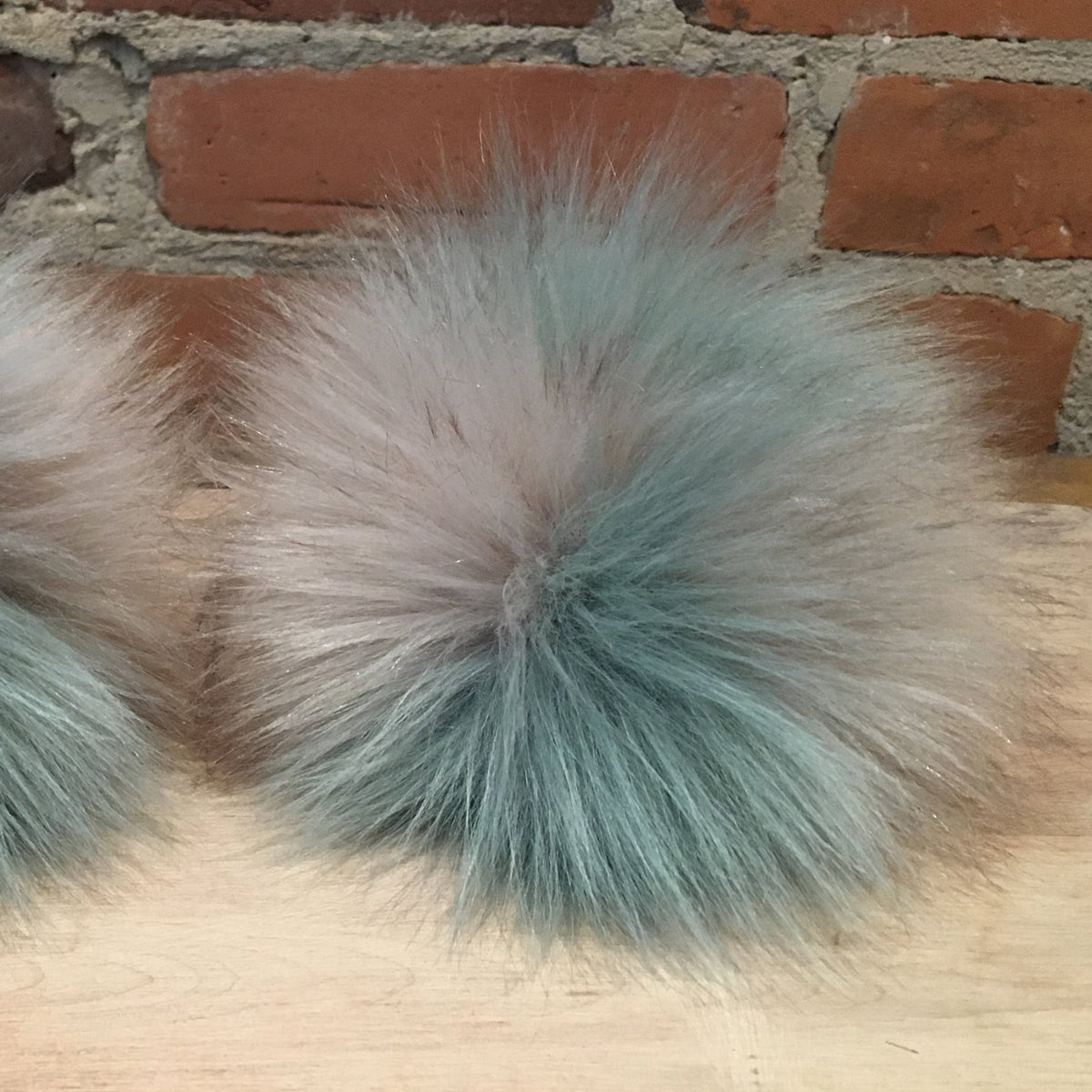 Two fluffy faux-fur pom-poms in a mottled gray and dusty blue-gray color, displayed side-by-side on a light wood surface with a red brick wall background.